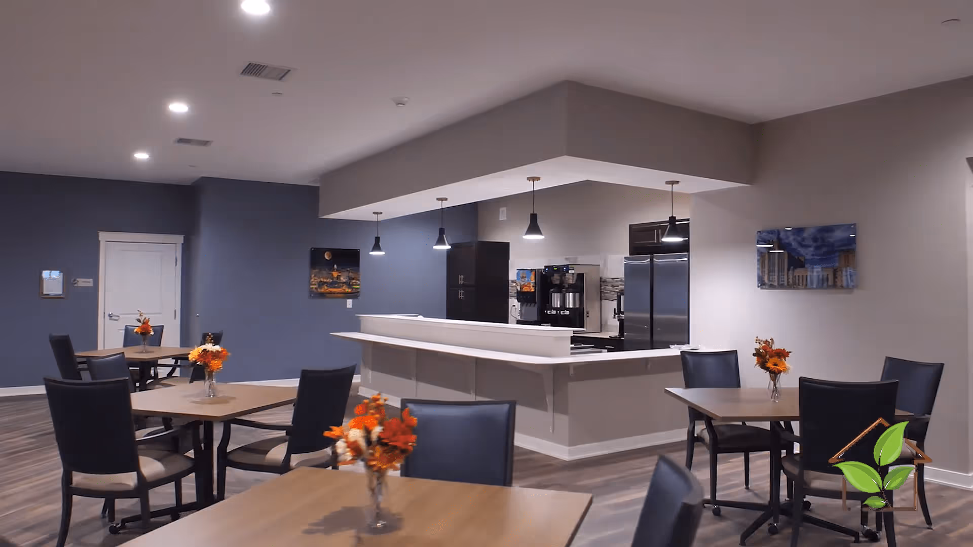 A modern dining area with several square wooden tables, each adorned with a small vase of orange and red flowers. The room features dark blue and beige walls, recessed ceiling lights, and a kitchen area with stainless steel appliances and hanging pendant lights in the background.