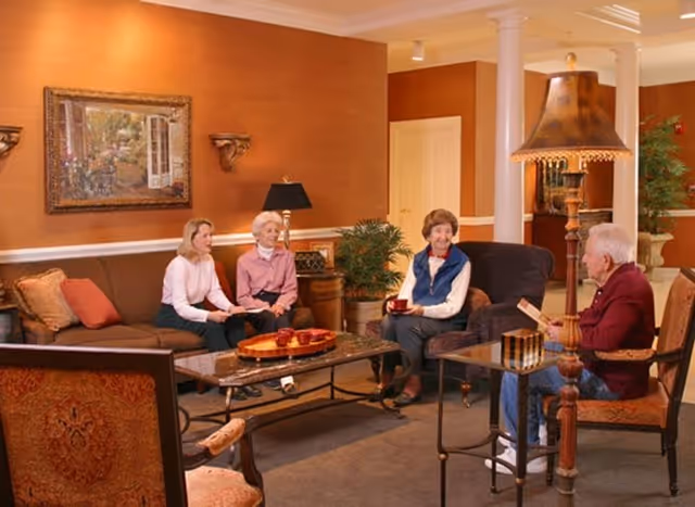 A cozy living room with warm lighting featuring four people sitting and conversing. There are two women seated on a brown couch, one woman on a dark armchair holding a cup, and a man sitting on a chair holding a book. The room has a framed painting on the wall, a table with decorative items, a large lamp, and some indoor plants.