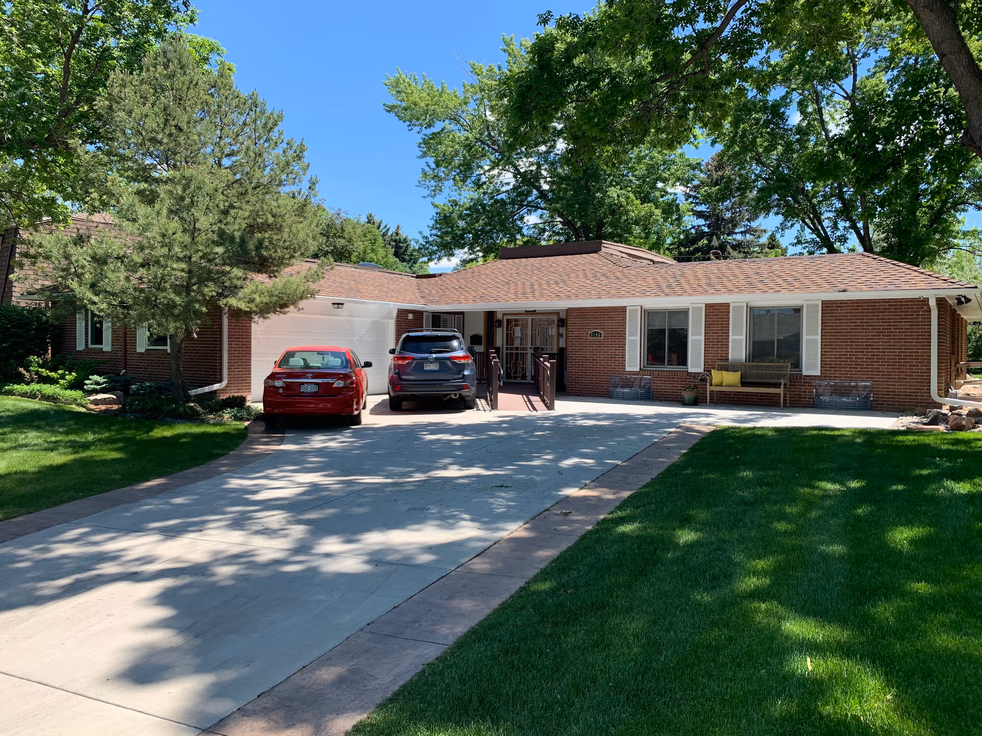 Front exterior of a single-story brick house with a driveway, two parked cars, and a shaded lawn with trees.