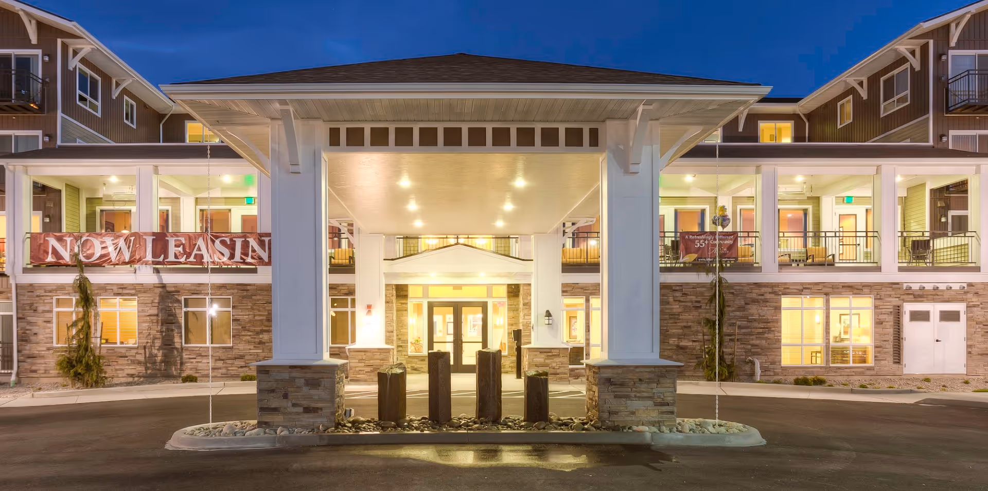 Entrance of a multi-story senior living building at dusk with a covered drop-off canopy, illuminated interior, and a 'NOW LEASING' banner.