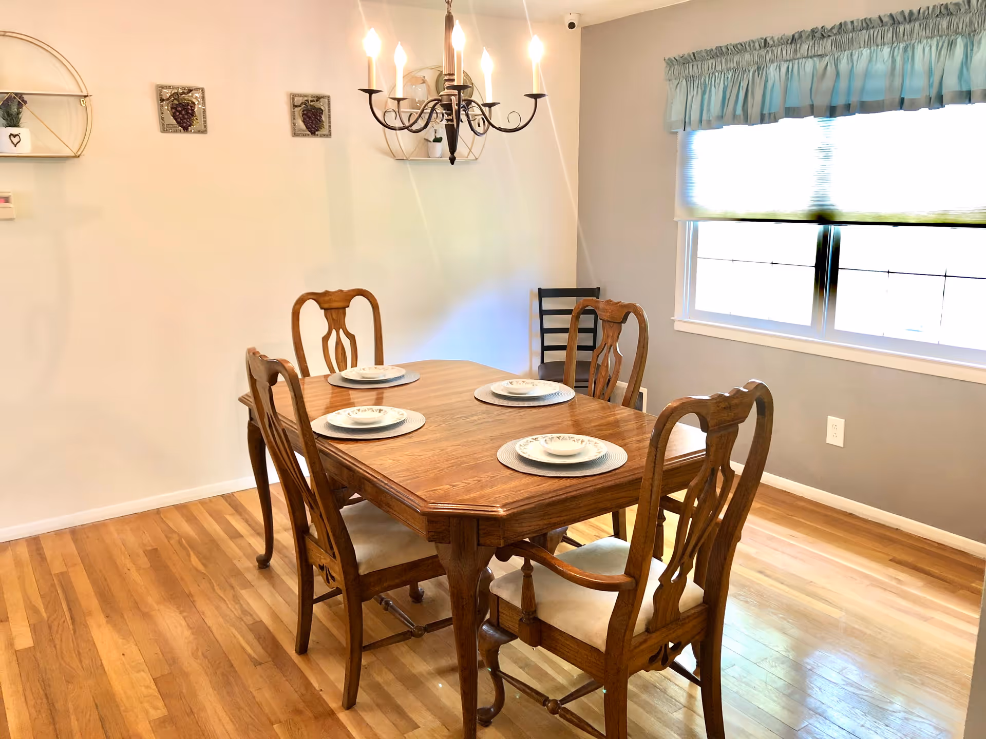 A dining room with a wooden table set for four with plates and bowls. Four wooden chairs surround the table, and a chandelier with lit bulbs hangs above. The room has hardwood floors, a window with a blue valance and a white blind, and light-colored walls decorated with small grape-themed wall hangings and shelves with small plants.