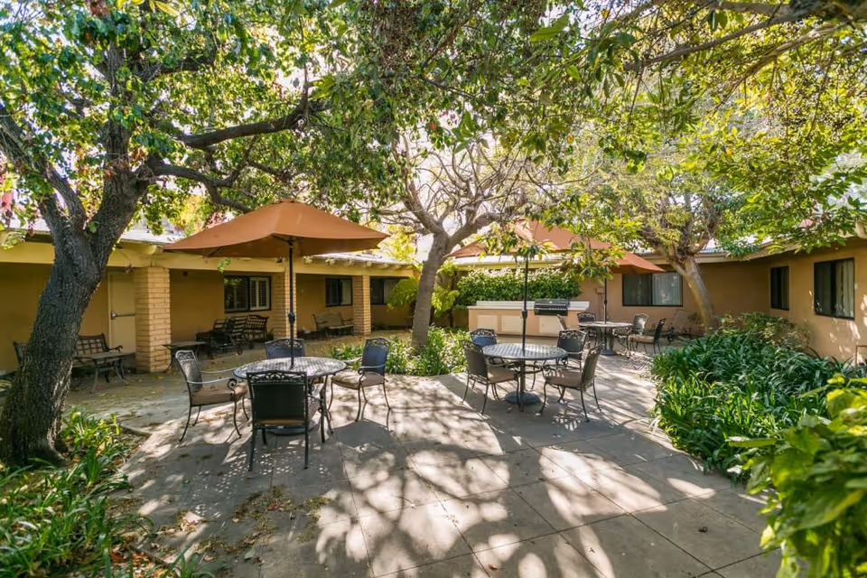 Outdoor courtyard area at Mainplace Senior Living with several round metal tables and chairs, each shaded by large brown umbrellas. The courtyard is surrounded by single-story building walls with windows and benches. Trees with green leaves provide additional shade and cast shadows on the paved ground. There are plants and greenery along the edges of the courtyard.