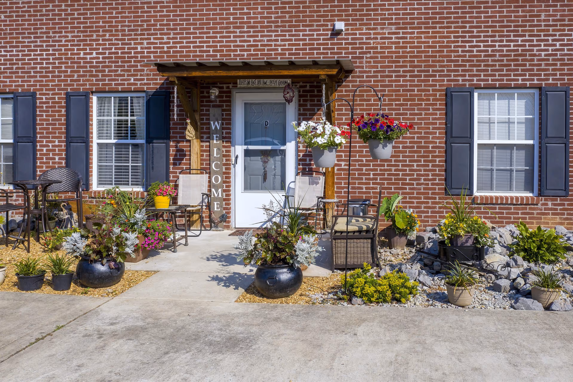 Entrance to a brick building with a white door labeled 'D' under a small wooden awning. There is a vertical wooden sign next to the door that says 'WELCOME'. The area is decorated with various potted plants and flowers, hanging flower pots, and outdoor seating including chairs and a small table. Two windows with black shutters flank the door.