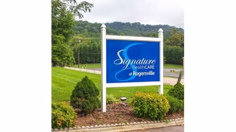 Outdoor blue sign that reads "Signature HealthCARE of Rogersville" mounted on white posts in a landscaped lawn with trees and hills in the background.
