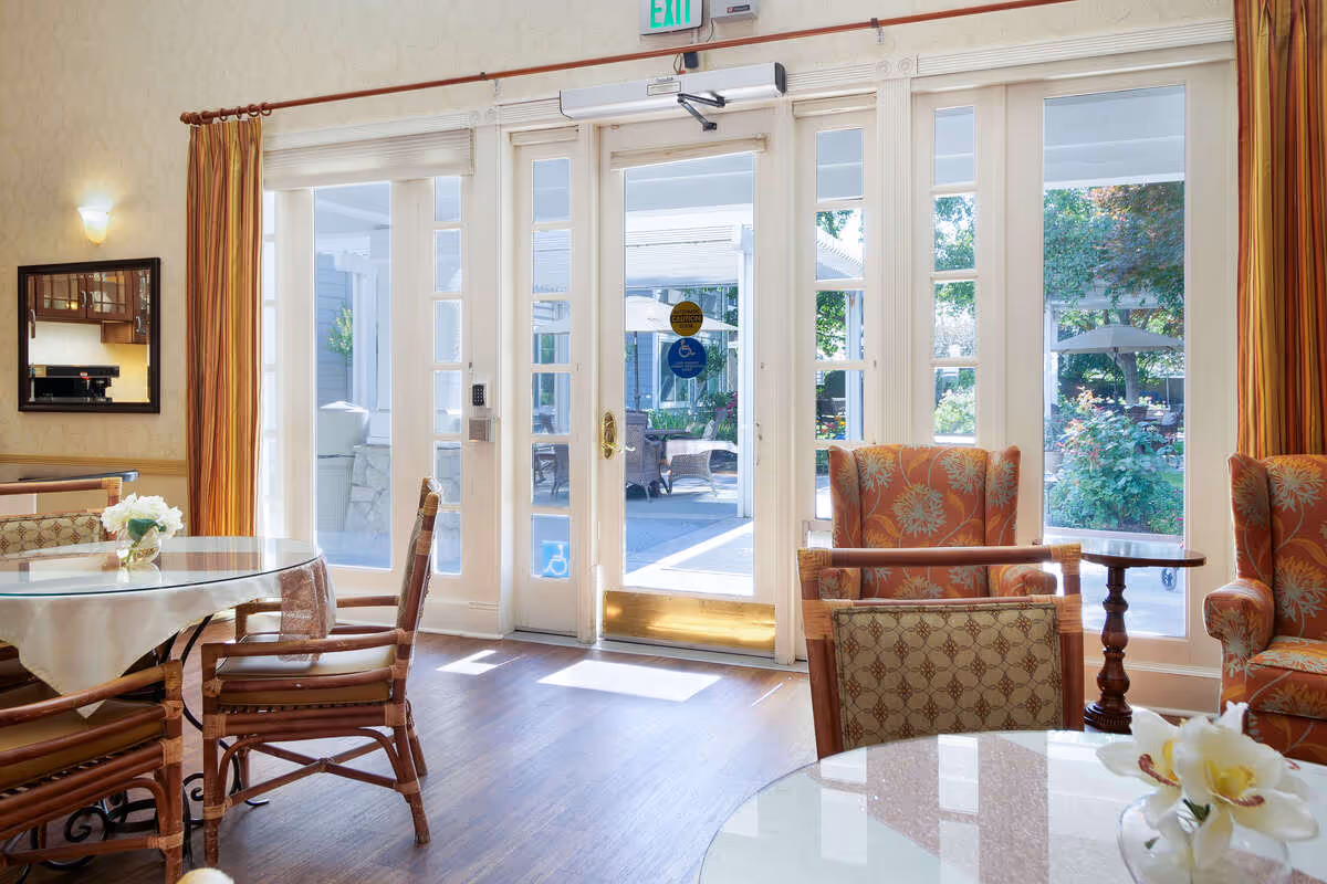 A bright and inviting sitting area in a senior living facility with glass double doors leading to an outdoor patio. The room features wooden floors, a round glass table with chairs, floral-patterned armchairs, and a small side table. Light filters in through the large windows, and there are curtains on either side of the doors.