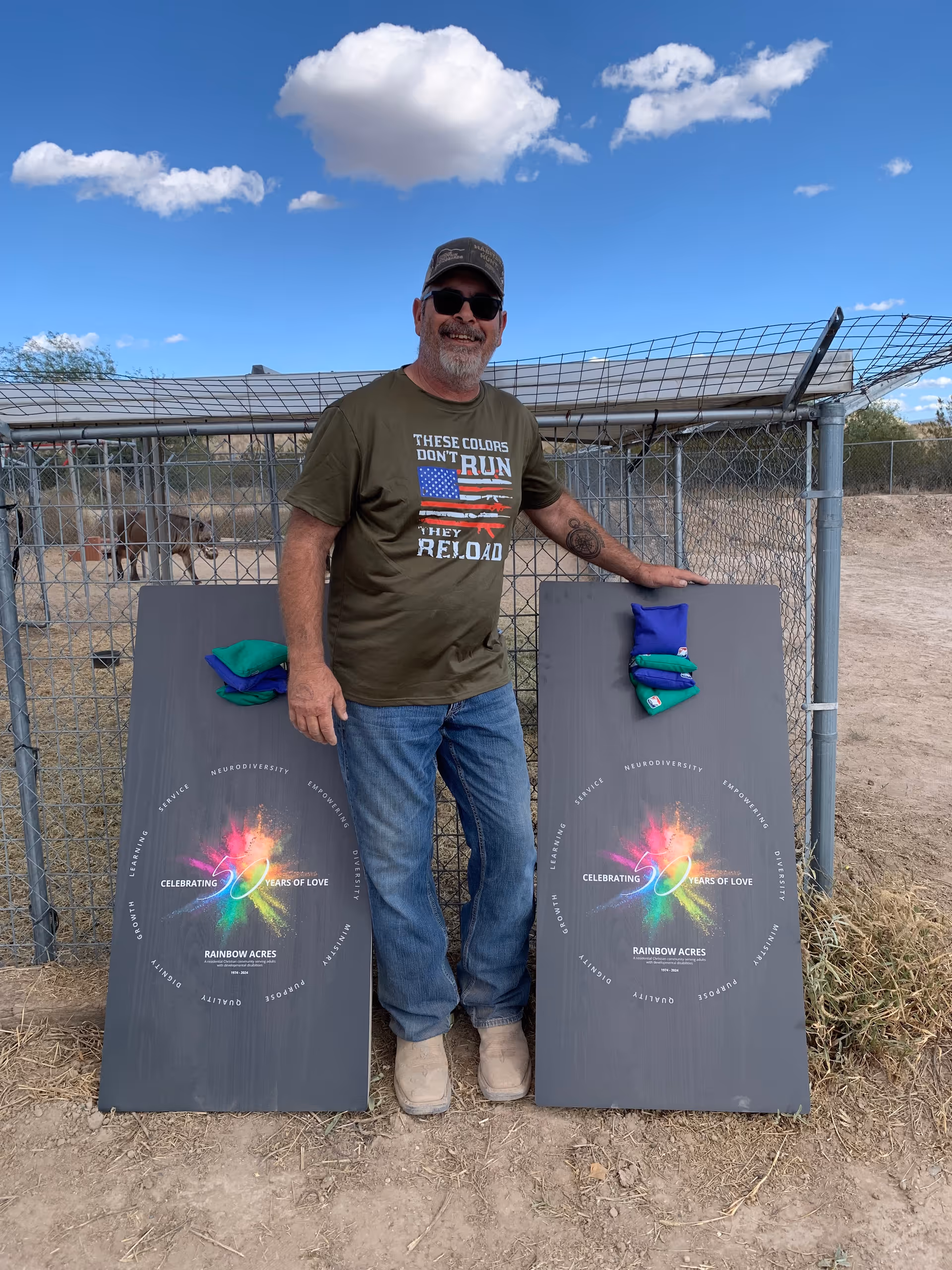 A man wearing sunglasses, a cap, a green t-shirt with an American flag design, and jeans is standing outdoors on dirt ground. He is smiling and posing between two cornhole boards that have colorful designs and the text 'Celebrating 70 Years of Love Rainbow Acres' printed on them. Behind him is a chain-link fence and a clear blue sky with a few clouds.