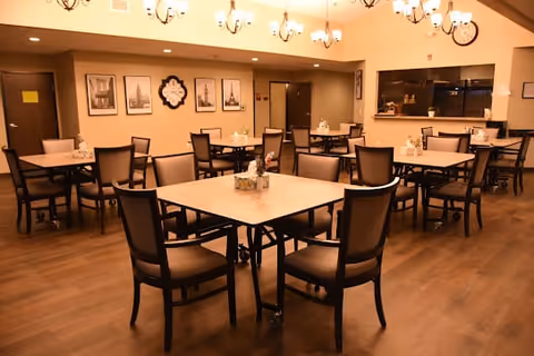 Well-lit dining room with multiple square tables and chairs, chandeliers overhead, and a serving counter in the back.