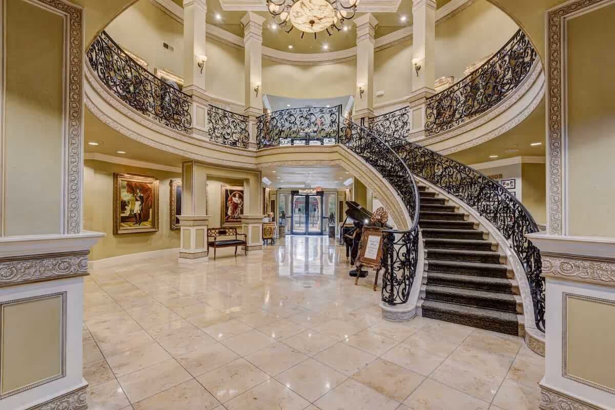 Spacious ornate lobby with a sweeping curved staircase, decorative wrought-iron railings, chandelier, marble floors and framed artwork.