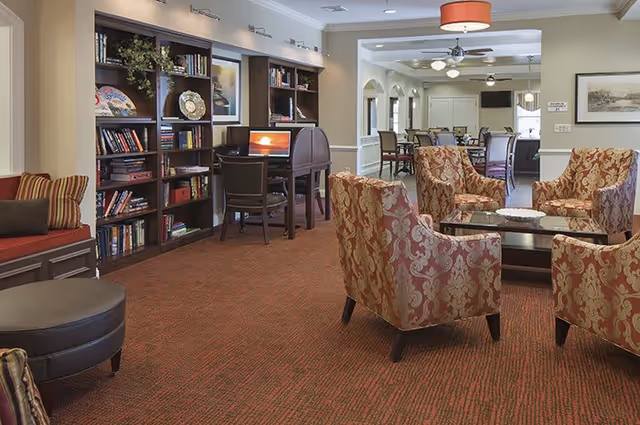 A cozy senior living common area with patterned armchairs arranged around a glass coffee table on a red carpet. There is a built-in bookshelf with books and decorative items, and a desk with a computer. In the background, there is a dining area with tables and chairs under ceiling fans and pendant lights.