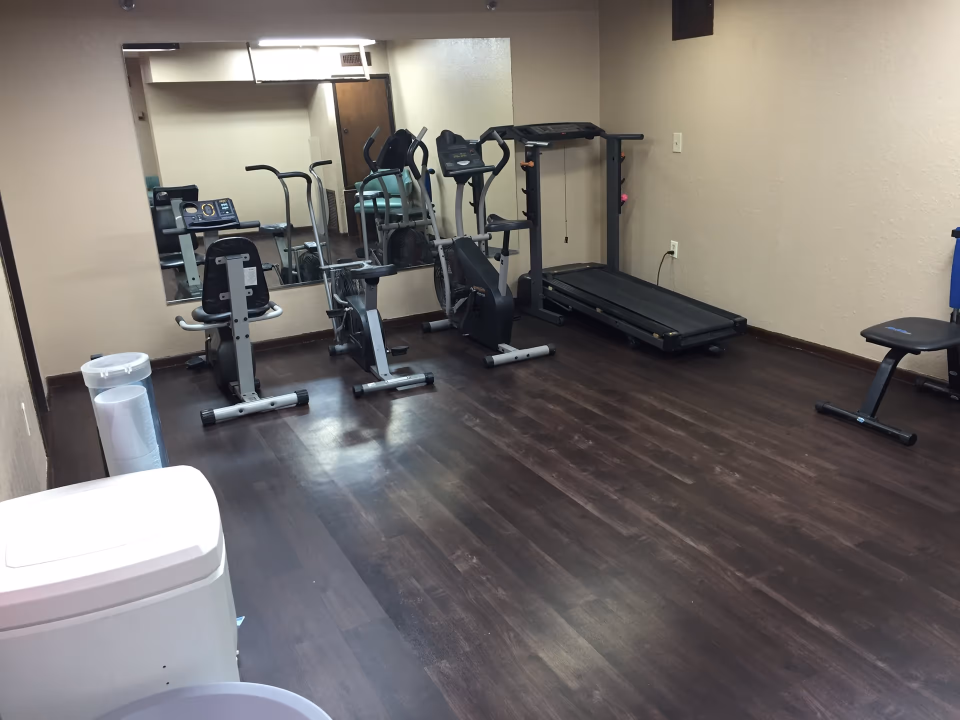 Small fitness room with a treadmill, stationary bikes, and an exercise bench in front of a wall mirror.