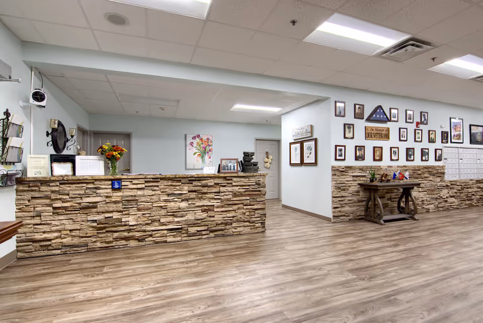 Reception area with a stone facade counter, wood flooring, and light blue walls. The wall behind the counter features framed pictures and a floral painting. To the right, there is a table with small flags and more framed photos on the wall above a row of mailboxes.