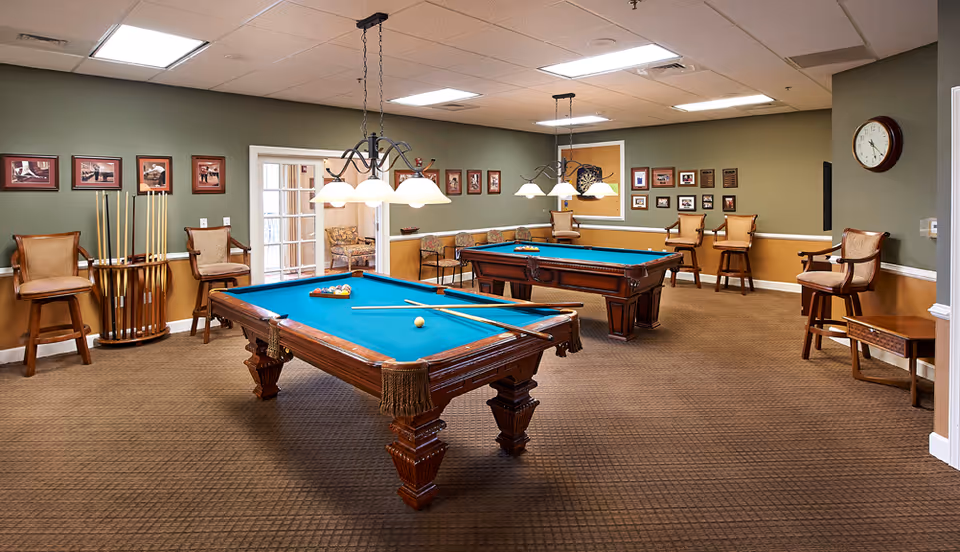 A recreational room with two pool tables, each with pool cues and balls set up. The room has green and beige walls decorated with framed pictures and plaques. Several wooden chairs with cushioned seats are arranged around the room, and two hanging light fixtures illuminate the pool tables. A clock is mounted on the wall to the right.