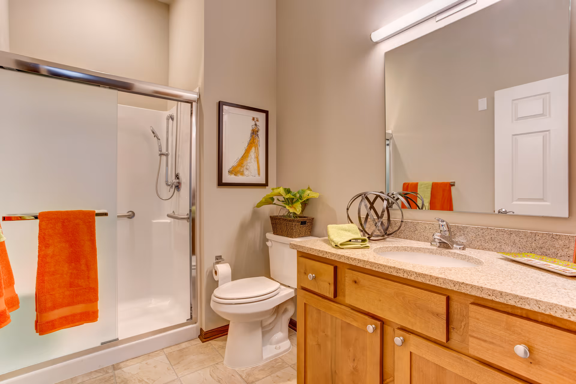 Bright bathroom with a glass sliding shower, toilet, wooden vanity with sink and large mirror, and orange towels.