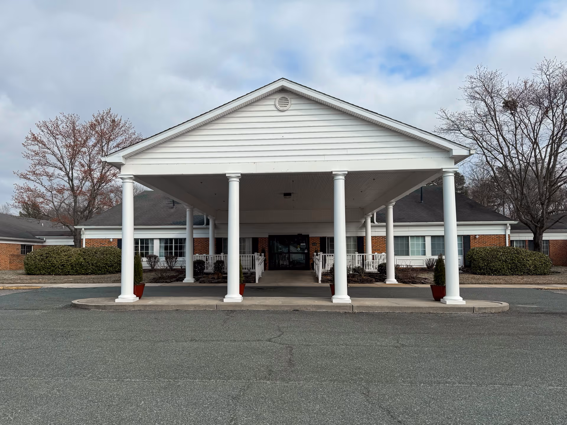 Front exterior view of a single-story brick building with a large white covered entrance supported by white columns. There are bushes and leafless trees on either side of the entrance, and the sky is partly cloudy.