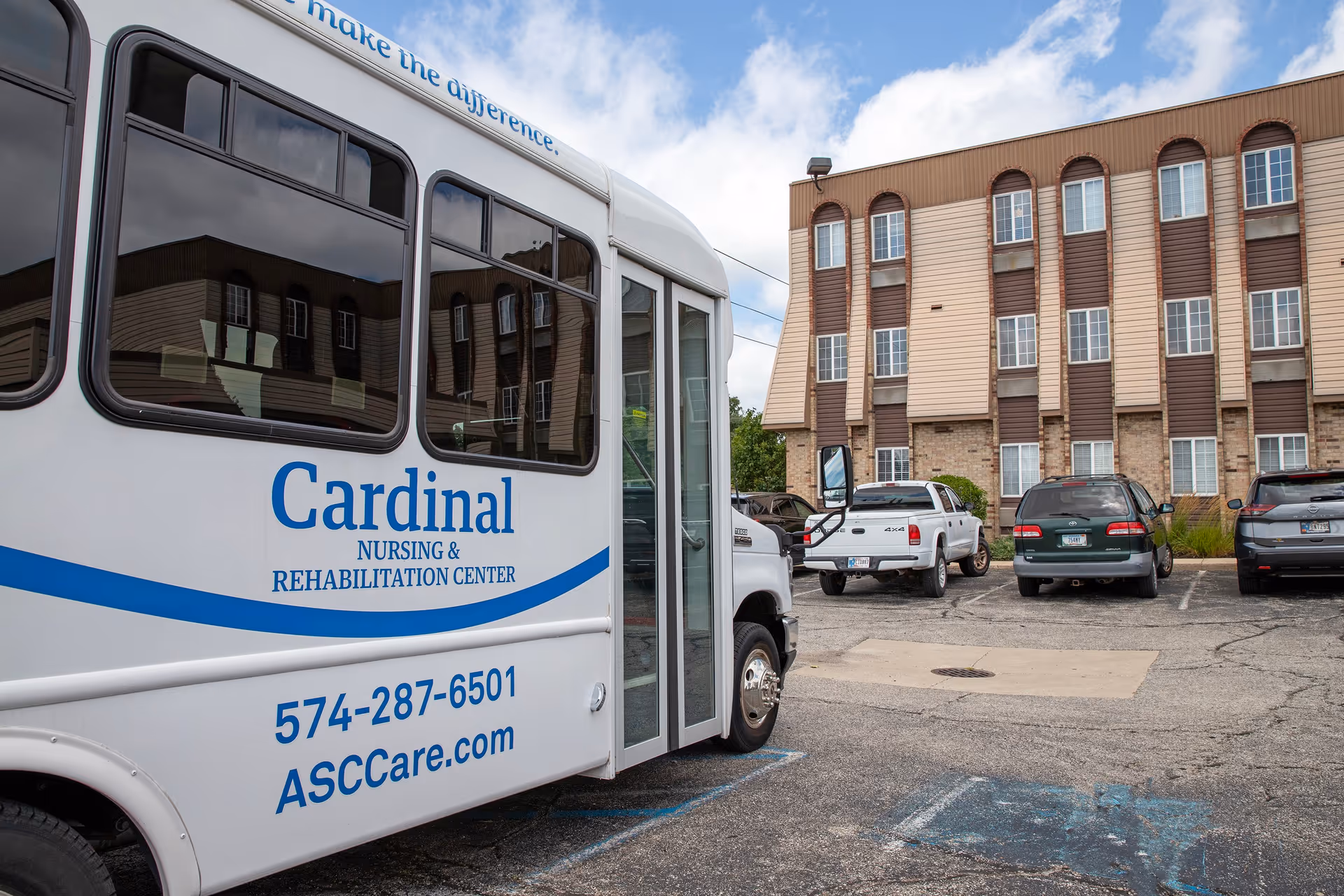 A white shuttle bus with the text 'Cardinal Nursing & Rehabilitation Center' parked in a parking lot in front of a multi-story building with several windows. Several cars are also parked in the lot under a partly cloudy sky.