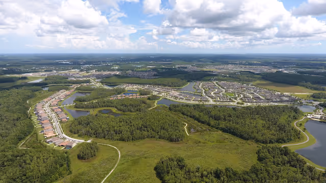 Aerial view of a residential community with rows of houses, lakes, and surrounding wetlands and forest under a partly cloudy sky.