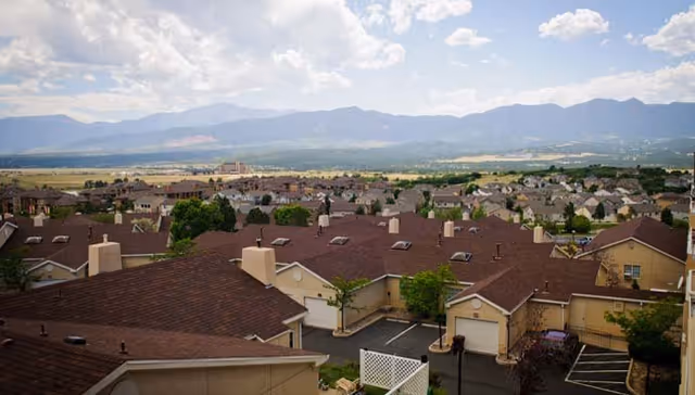 View over a neighborhood of tan-roofed residential buildings with mountains and a cloudy sky in the background.