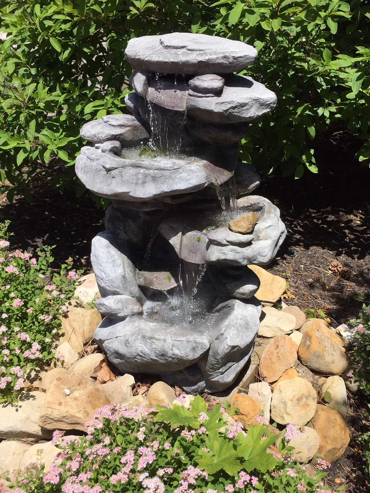 A multi-tiered stone water fountain with water trickling into rock basins surrounded by pink flowers and green foliage.
