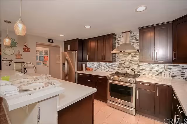 Modern kitchen with dark wood cabinets, stainless steel appliances including a refrigerator and stove with a range hood, a tiled backsplash, and a white countertop island with pendant lights above.