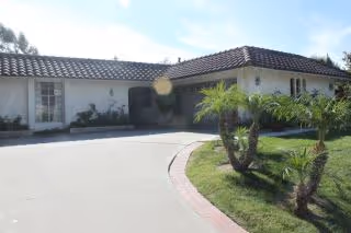 Exterior view of a single-story building with a tiled roof, white walls, and several windows. There is a curved driveway leading to the entrance, and a small landscaped area with palm trees and grass on the right side.