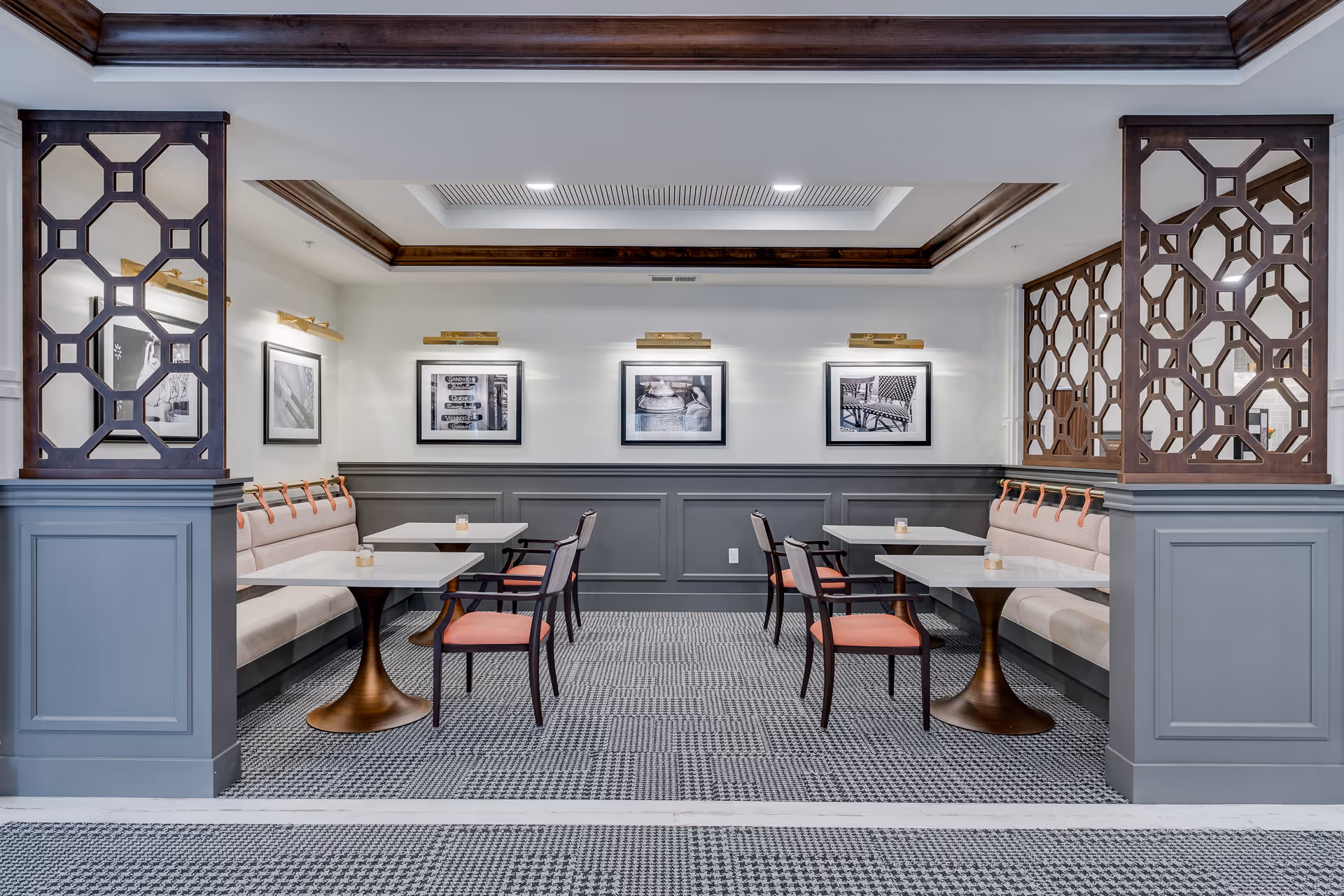 A dining area with two sets of tables and chairs arranged opposite each other. Each side has a cushioned bench with beige upholstery and wooden chairs with coral-colored seats. The walls are painted gray on the lower half and white on the upper half, decorated with three black and white framed photographs. The ceiling has recessed lighting and wooden crown molding, and decorative wooden lattice panels partially separate the dining space from the surrounding area.