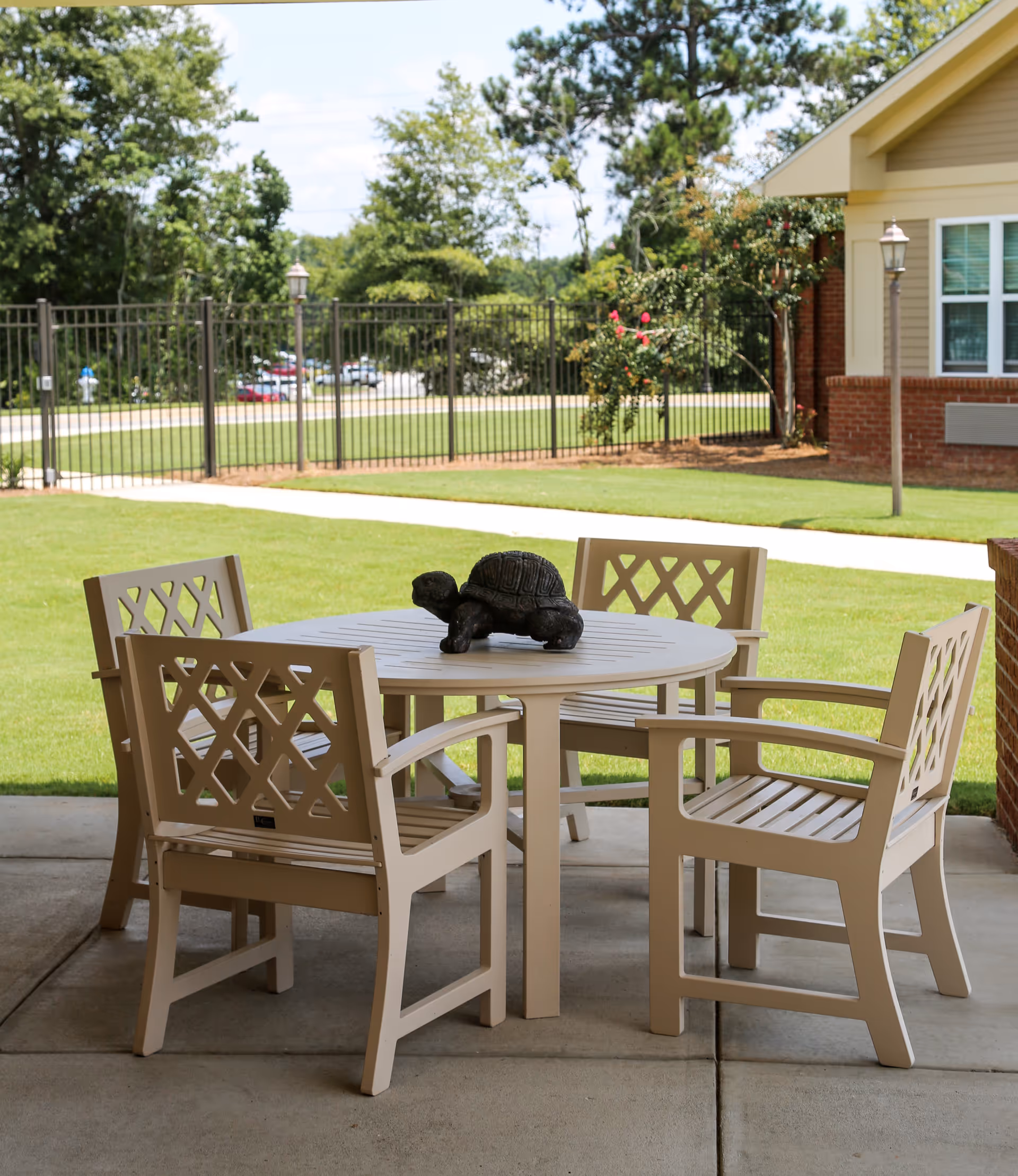 Outdoor patio area with a round beige table and four matching chairs. A decorative turtle sculpture is placed on the table. The patio overlooks a well-maintained lawn with a black metal fence and trees in the background. Part of a building with brick and beige siding is visible on the right side.