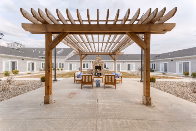 Outdoor courtyard area at Cedarhurst Senior Living of Fort Wayne featuring a wooden pergola with seating underneath, including wooden chairs with cushions and a central stone fireplace. The courtyard is surrounded by single-story buildings with white siding and multiple windows and doors.