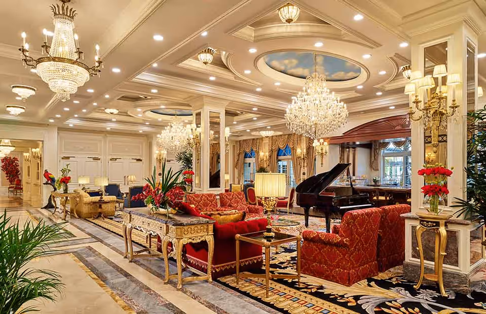 Lavish common lounge with crystal chandeliers, a grand piano, red upholstered sofas, ornate tables, and decorative plants.