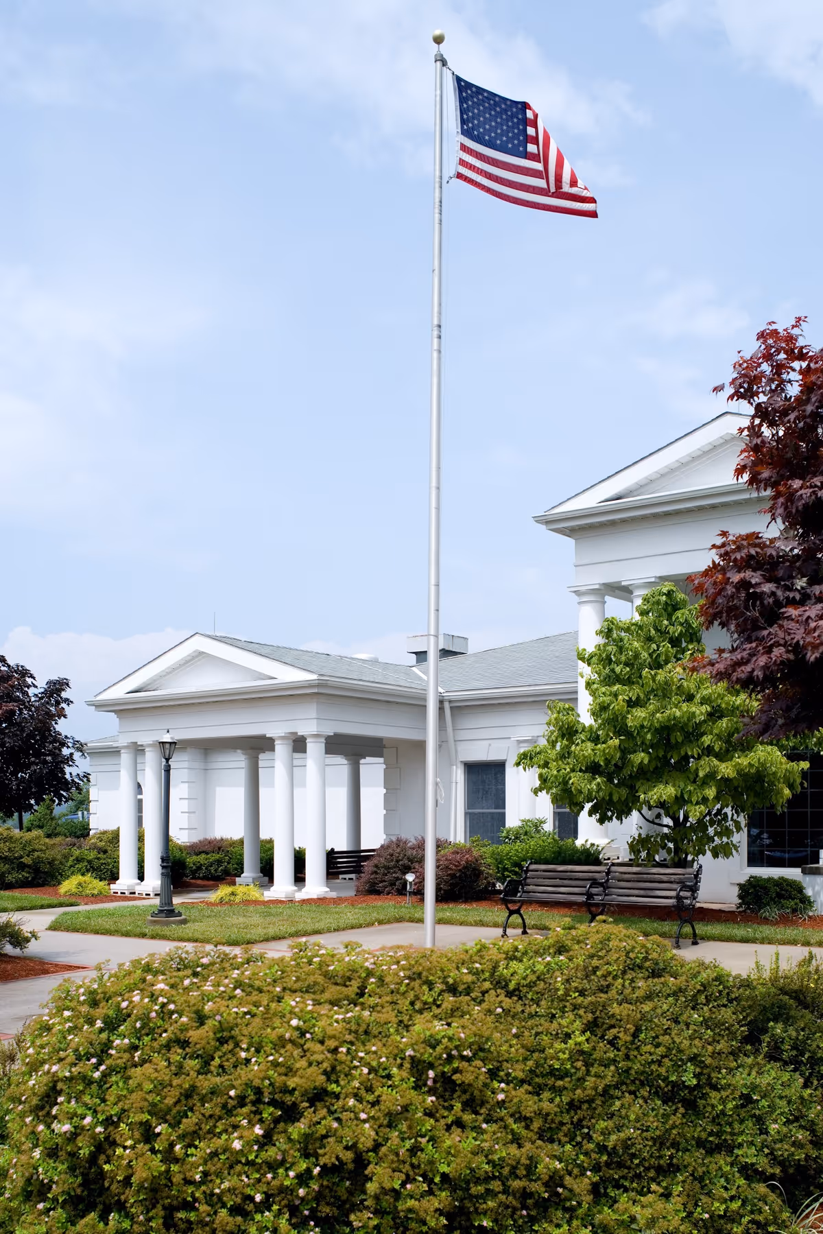 Front entrance of a white, columned building with an American flag on a tall flagpole, benches, and landscaped shrubs.