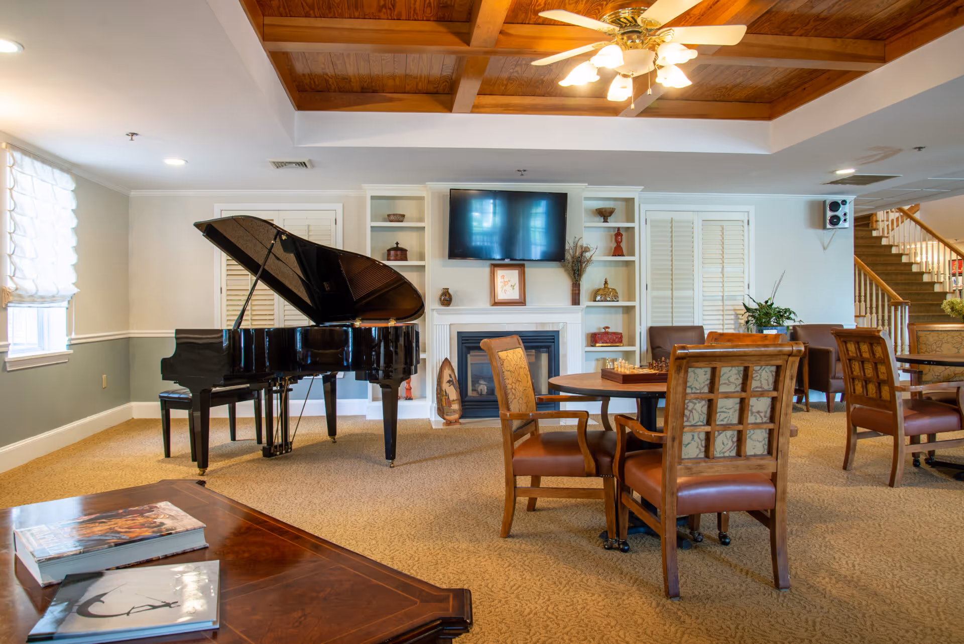 Spacious senior living common room with a grand piano, seating areas, fireplace, TV, and wood-paneled ceiling with a fan.