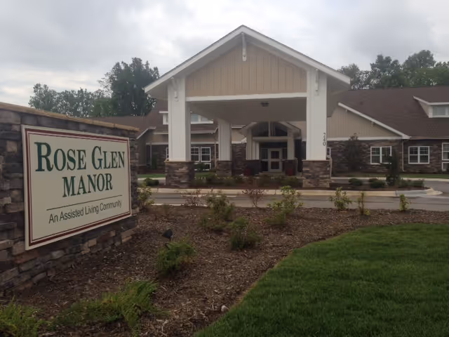 Exterior view of Rose Glen Manor, an assisted living community, showing the main entrance with a covered drop-off area, stone and wood facade, and landscaped grounds with grass and small shrubs.