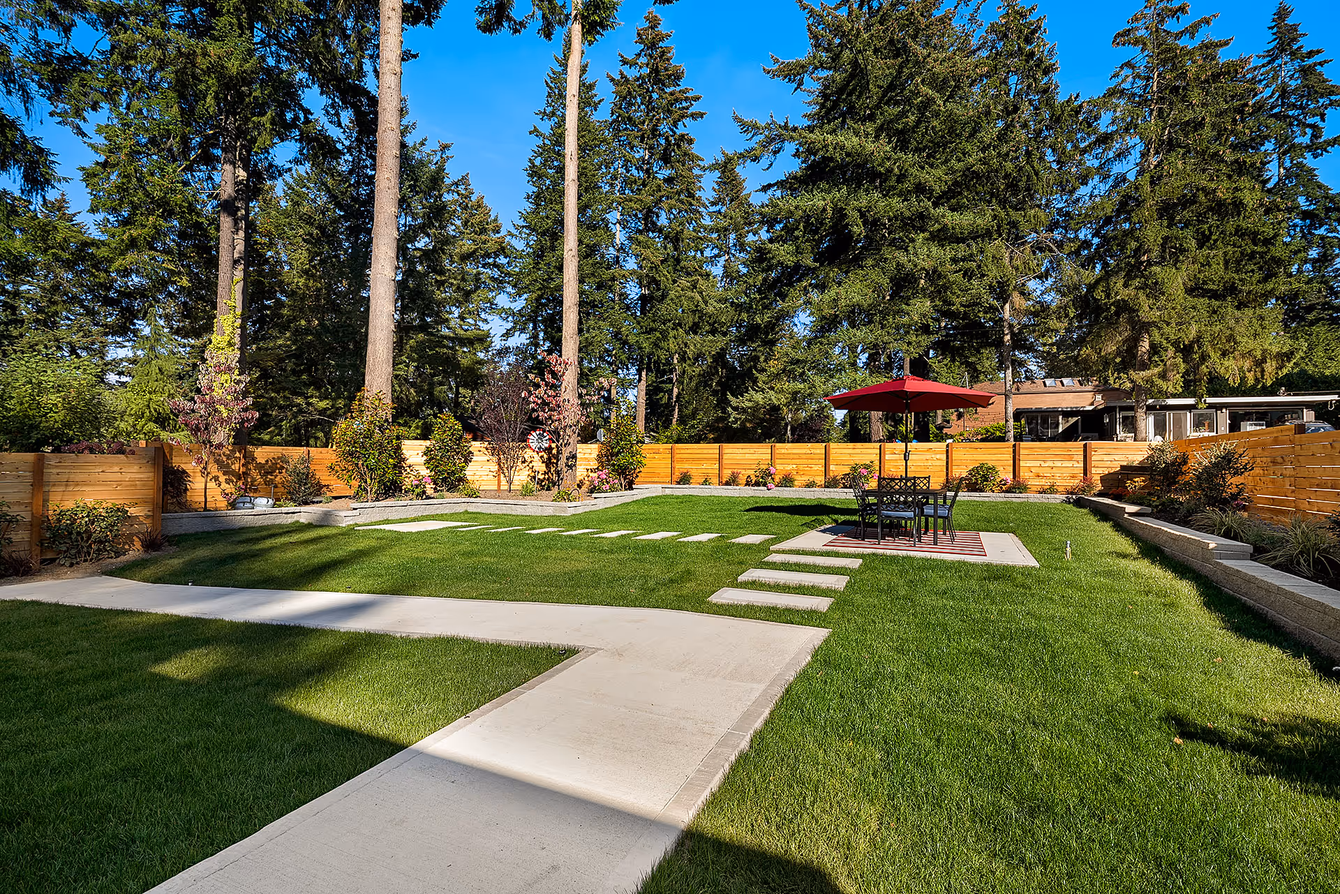 A well-maintained outdoor garden area with green grass, a concrete pathway, and a seating area with a table and chairs under a red umbrella. The garden is enclosed by a wooden fence and surrounded by tall trees under a clear blue sky.