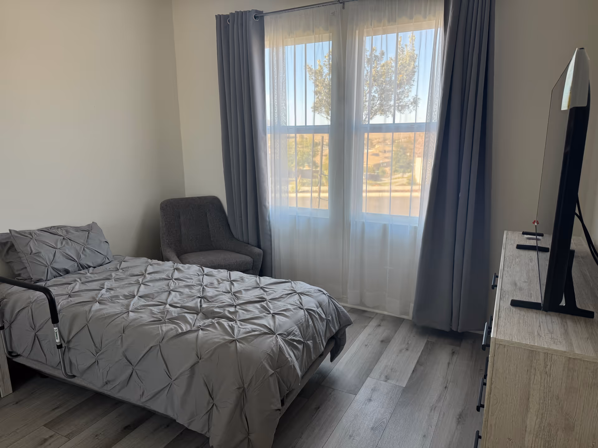 A bedroom with a single bed covered in a gray textured comforter, a gray upholstered chair in the corner, a large window with sheer white curtains and gray drapes, and a wooden dresser with a flat-screen TV on top.