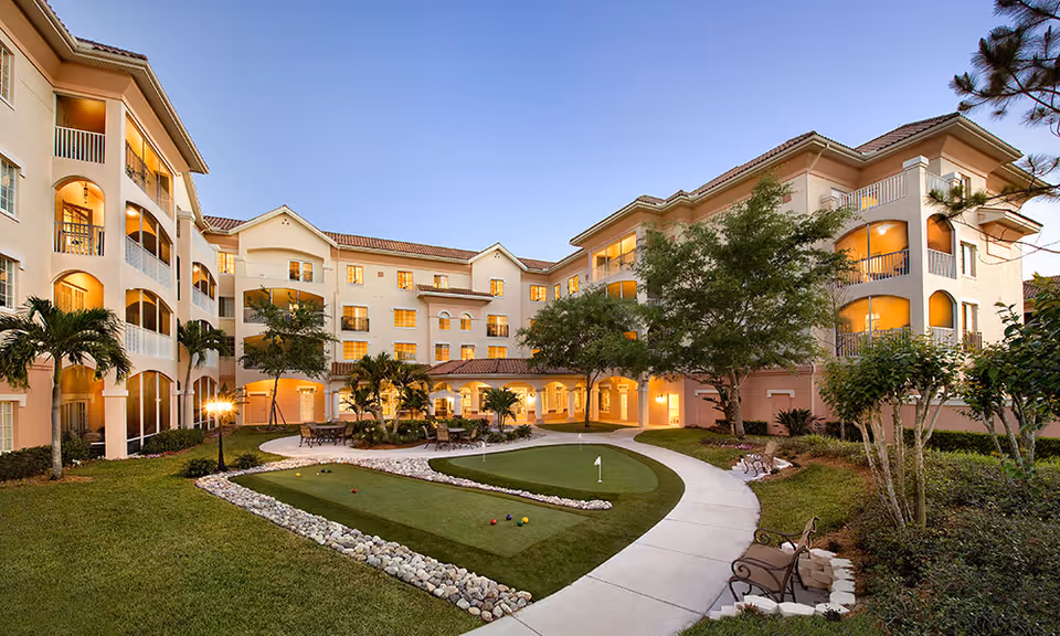 Outdoor courtyard area of Terracina Senior Living featuring a putting green, curved walking path, benches, palm trees, and a multi-story residential building with balconies and warm lighting at dusk.