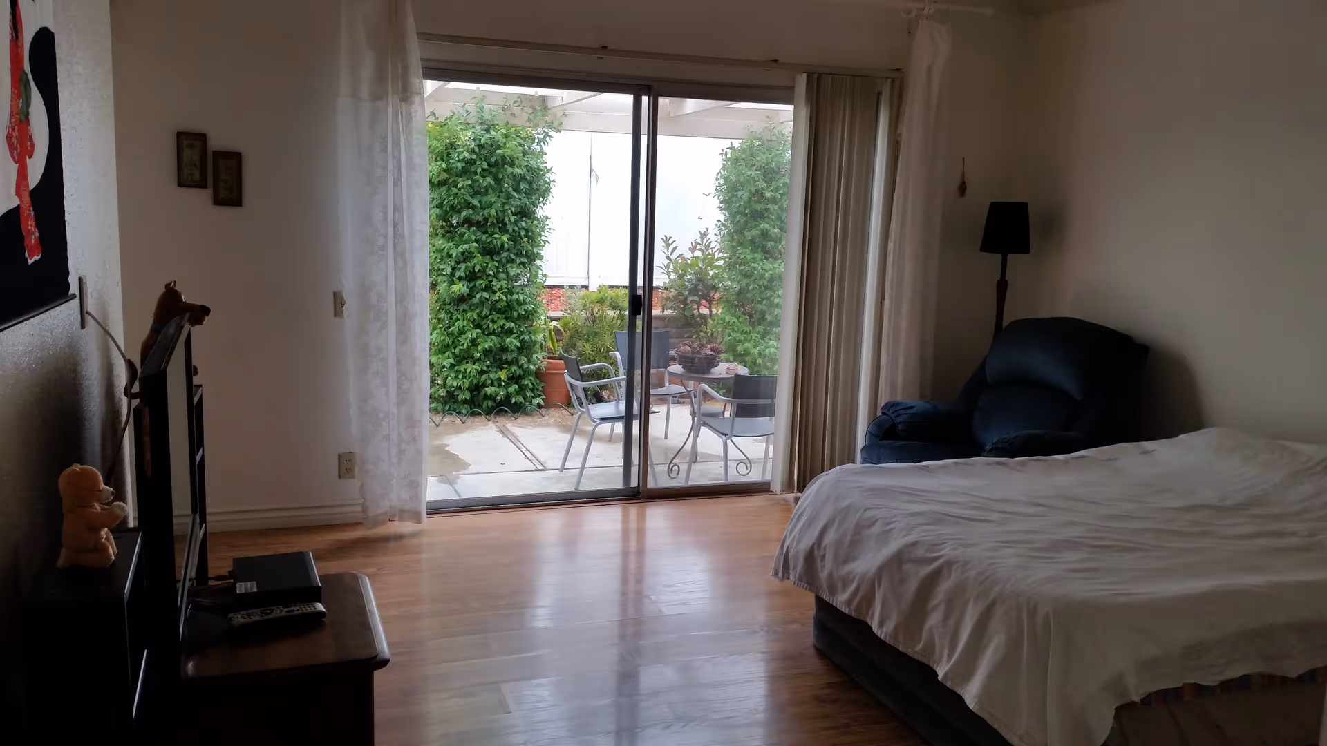 A bedroom with a bed covered in white sheets, a dark blue armchair, a floor lamp, and a TV on a stand with stuffed animals. The room has wooden flooring and a large sliding glass door leading to an outdoor patio with chairs and greenery.