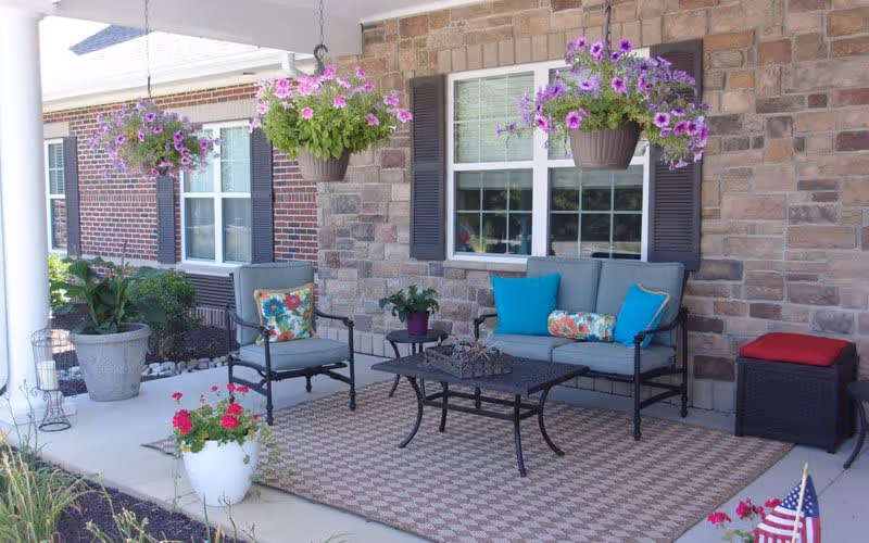A cozy outdoor patio area with cushioned seating including a loveseat and a chair with colorful pillows. There is a black metal coffee table on a patterned outdoor rug. Hanging flower pots with purple flowers are suspended from the ceiling. The background features a brick and stone wall with windows and shutters. Various potted plants and flowers decorate the space.
