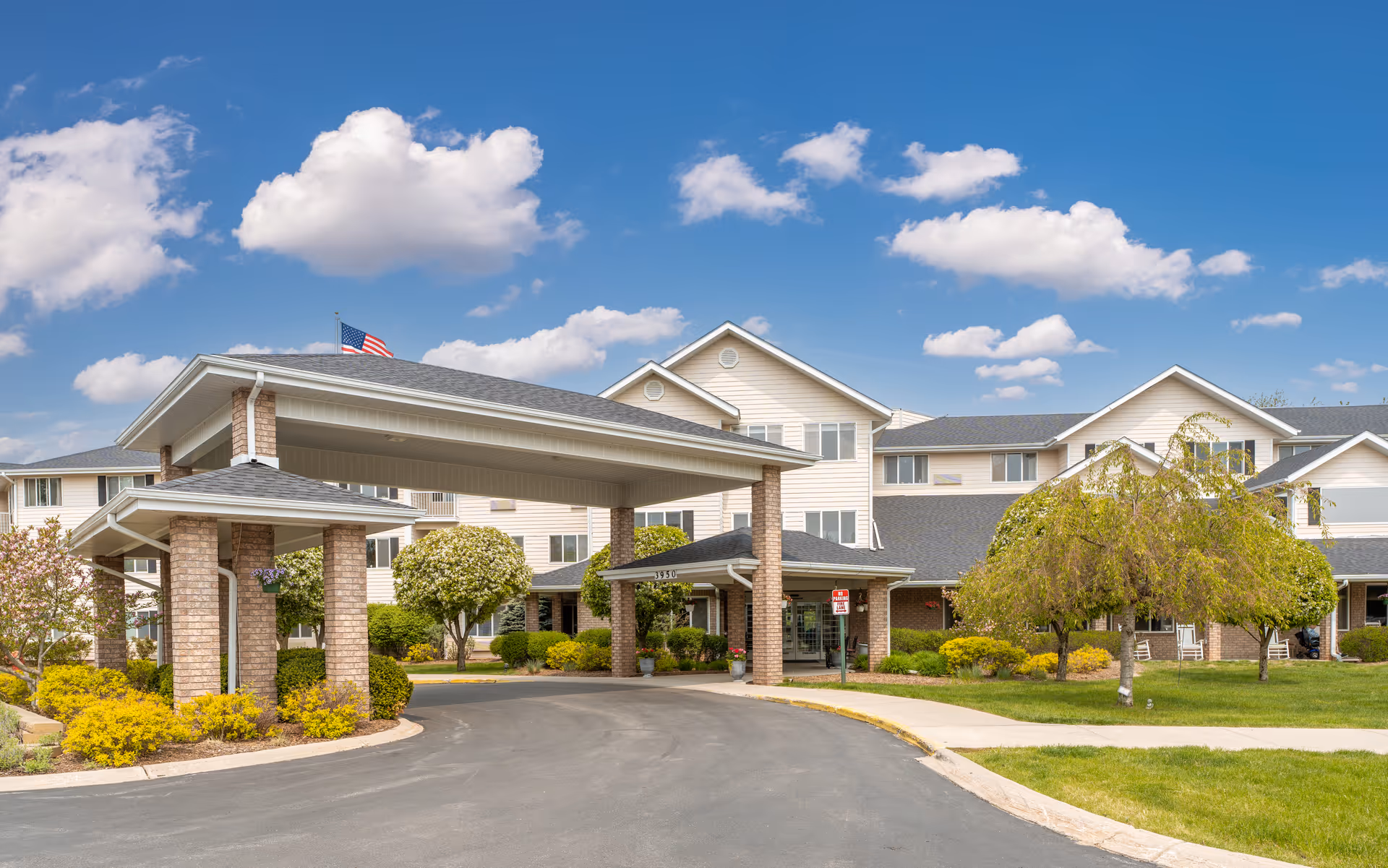 Exterior view of Independence Village of Columbus, showing a large covered entrance with brick columns, a paved driveway, well-maintained landscaping with bushes and trees, and a multi-story building under a blue sky with scattered clouds.