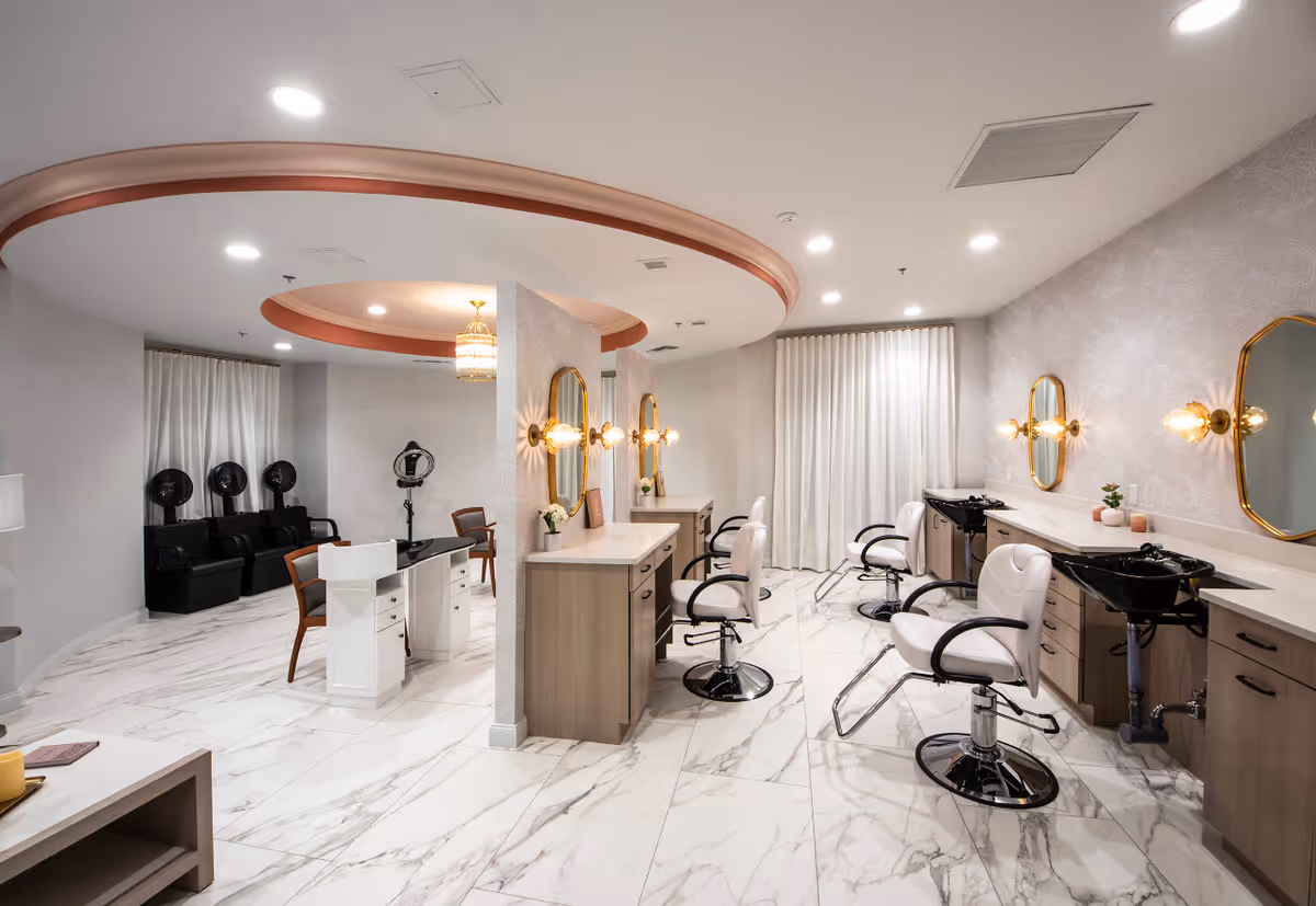 A modern salon area inside a senior living facility with multiple styling stations featuring mirrors, chairs, and sinks. The room has marble-patterned flooring, soft lighting, and a circular ceiling design with a chandelier. There are also hair drying chairs in the background.