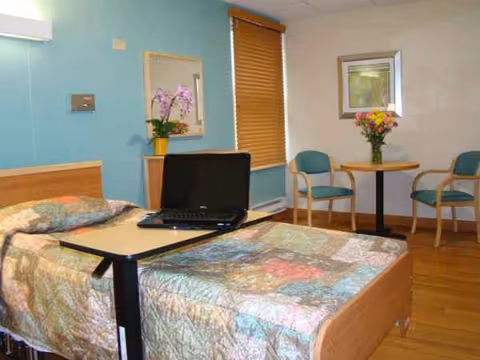 Bedroom in a senior facility with a bed, laptop on a table, and seating area.