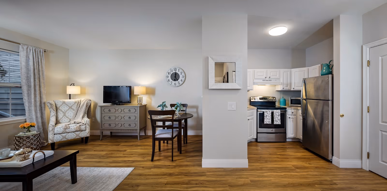 A cozy and well-lit living space featuring a sitting area with an armchair and side table, a small dining table with two chairs, and a compact kitchen with white cabinets, stainless steel appliances including a refrigerator and stove. The room has wooden flooring, a wall clock, two table lamps, and a window with curtains.