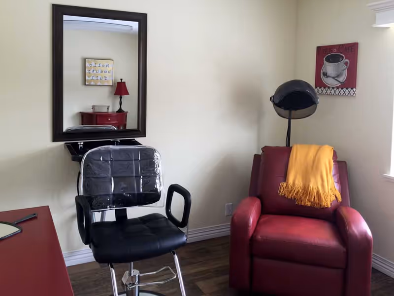 A small room with a black salon chair covered in plastic in front of a wall-mounted mirror and sink. To the right, there is a red leather recliner with a yellow throw blanket draped over it, and a vintage hair dryer hood stands behind the recliner. The walls are light-colored, and there is a small framed coffee-themed artwork on the wall near a window.