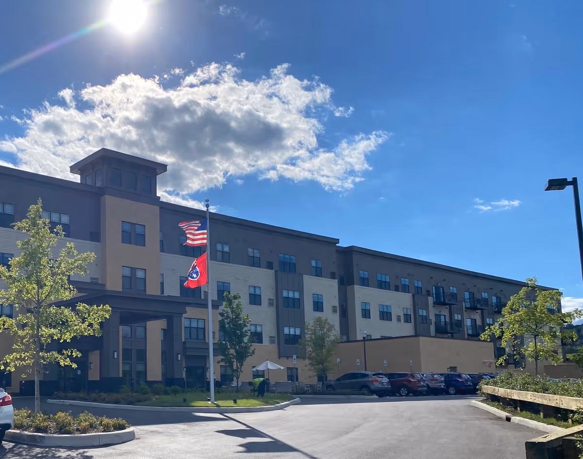 Exterior view of a multi-story senior living facility building under a bright blue sky with some clouds. There are two flagpoles in front of the building displaying the American flag and the Tennessee state flag. Several cars are parked in the parking lot, and small trees and landscaping are visible around the entrance.
