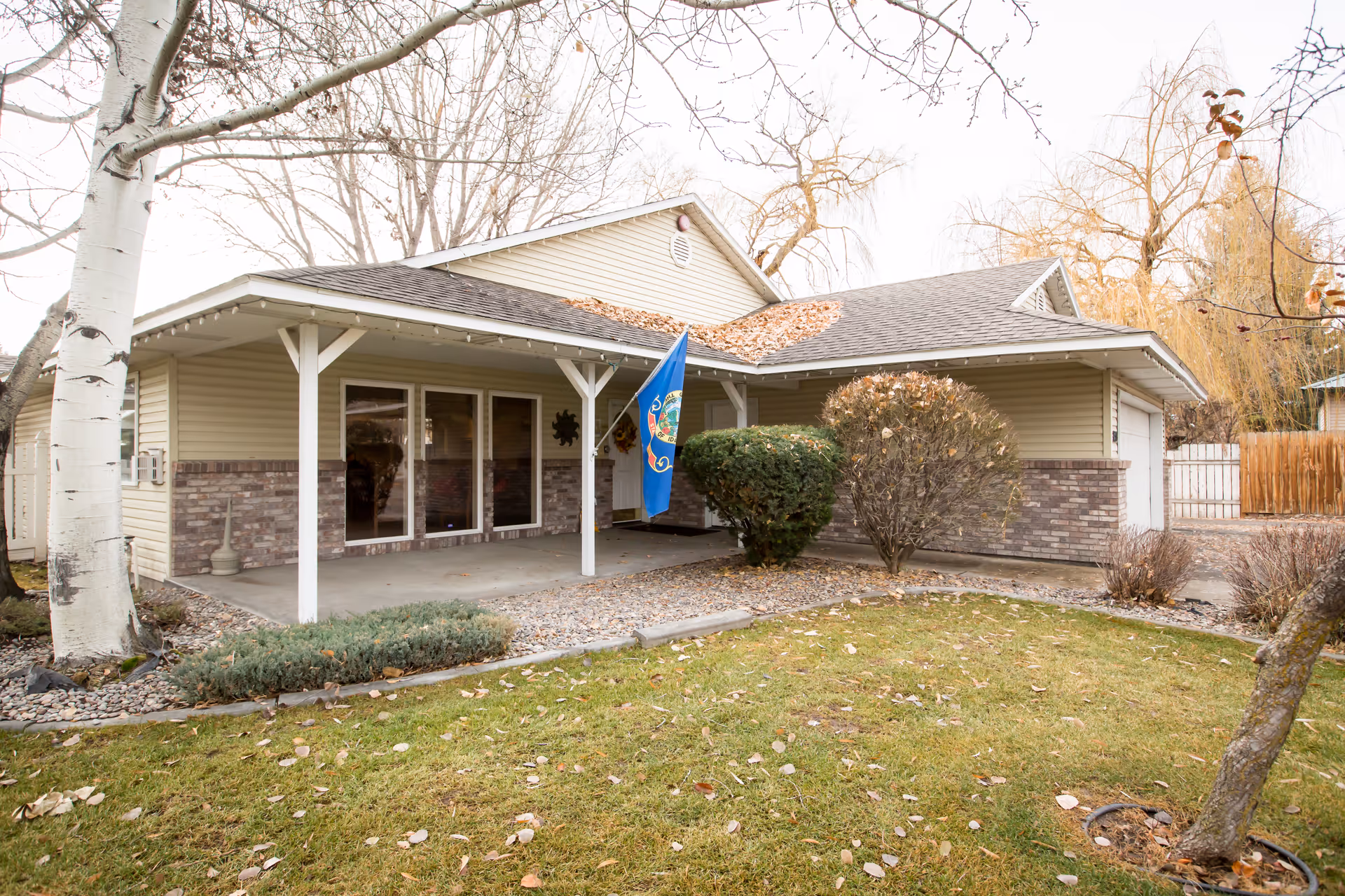 Front exterior of a single-story house with a covered porch, flag, shrubs, and a grassy yard.