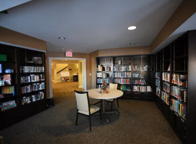 Interior view of a library or reading room with dark wooden bookshelves filled with books lining the walls. In the center, there is a round table with two chairs and a small decorative centerpiece. The room has carpeted flooring and a doorway leading to a well-lit hallway.