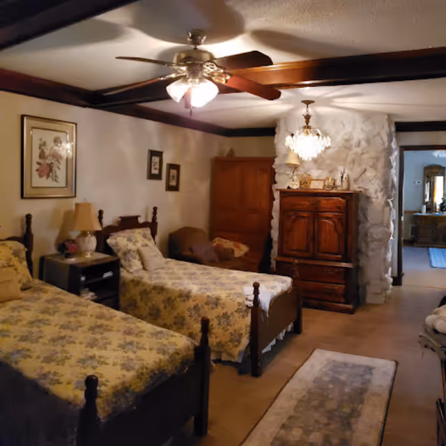 A cozy bedroom with two twin beds covered in floral bedspreads, a wooden nightstand with a lamp between the beds, framed floral artwork on the wall, a ceiling fan with lights, a wooden armoire against a white textured stone wall, and a small rug on the wooden floor.