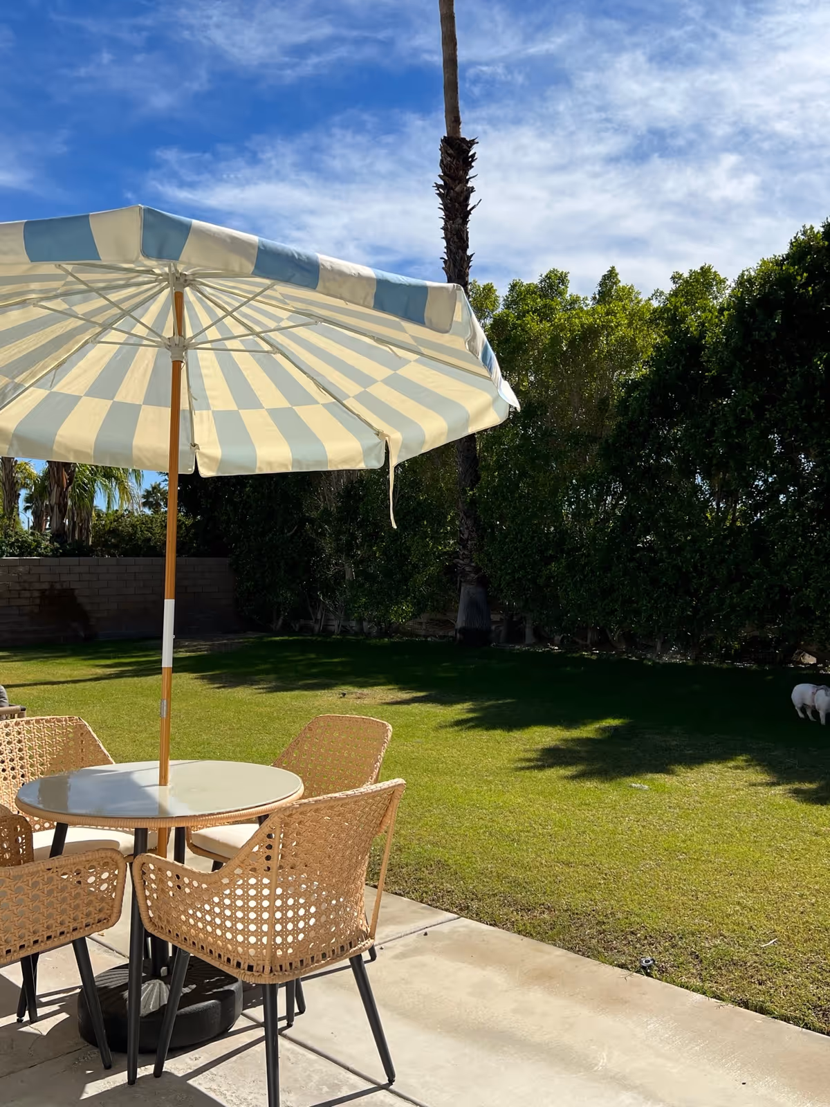 Outdoor patio area with a round table and four wicker chairs under a large blue and white striped umbrella. The patio overlooks a green lawn with trees and bushes in the background under a partly cloudy blue sky.