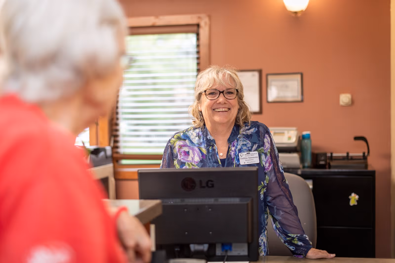 A smiling woman with glasses and a floral blouse stands behind a reception desk with a computer monitor. She is wearing a name tag that reads Debbie Simmons. Another person with white hair and a red shirt is seen in the foreground, slightly out of focus. The background shows a window with blinds and framed certificates on a peach-colored wall.