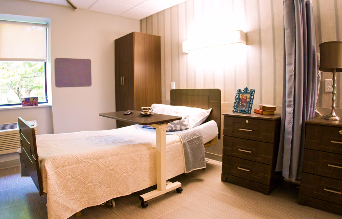 A neatly arranged bedroom in a senior living facility with a single bed covered in a white quilt, a bedside table with a lamp and framed photo, a wooden wardrobe, and a window with a roller shade partially down. A movable overbed table holds a cup and saucer and a remote control.