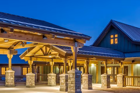 Exterior view of Mesa View Assisted Living facility at dusk, showing a covered walkway with wooden beams and stone pillars, and a building with green siding and a metal roof illuminated by warm lights.