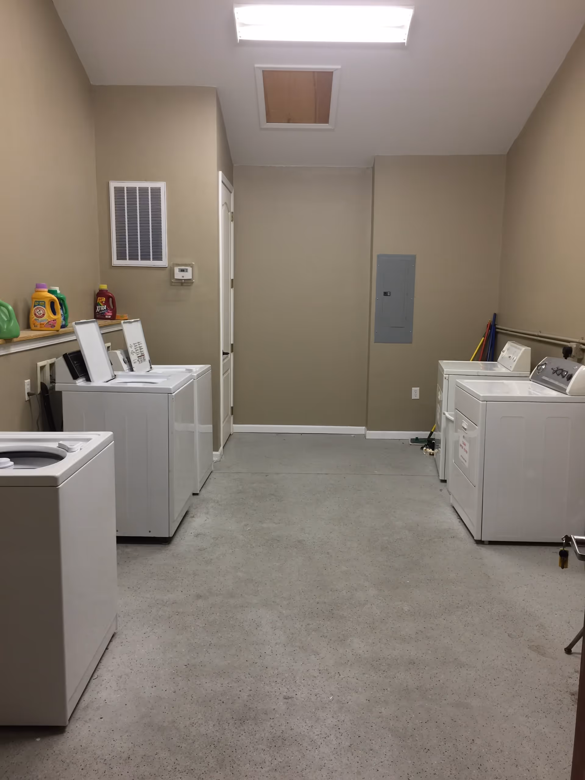 Laundry room with four white washing machines and dryers, laundry detergent bottles on a ledge, beige walls, and a gray speckled floor.